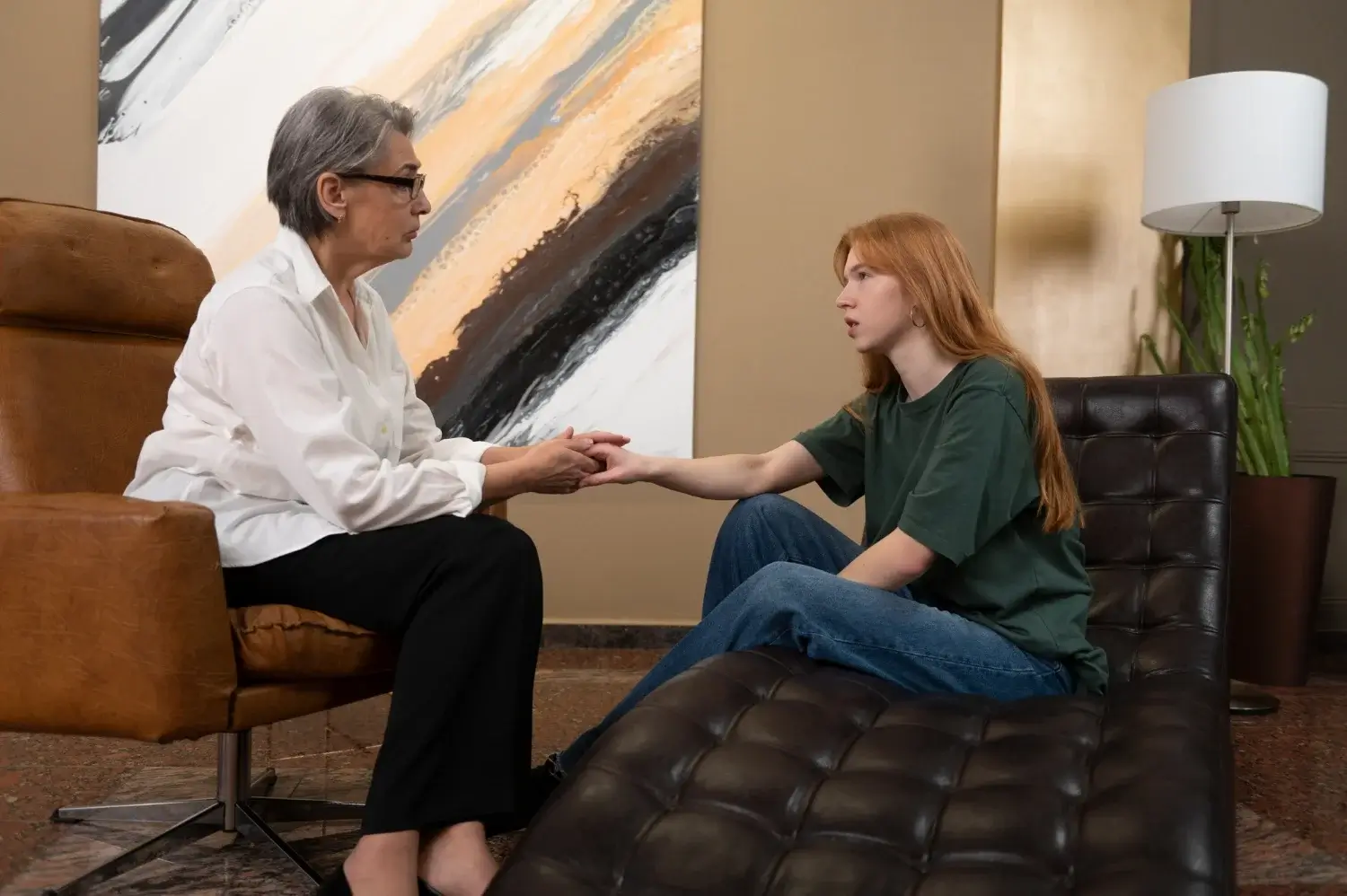 Coping with Life Transitions A Therapist’s Guide to Navigating Change A therapist, an older woman with grey hair, holds the hand of a younger woman with reddish-brown hair as they sit on a couch during a therapy session.